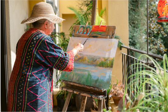 A woman in a hat and dress painting a landscape on a patio